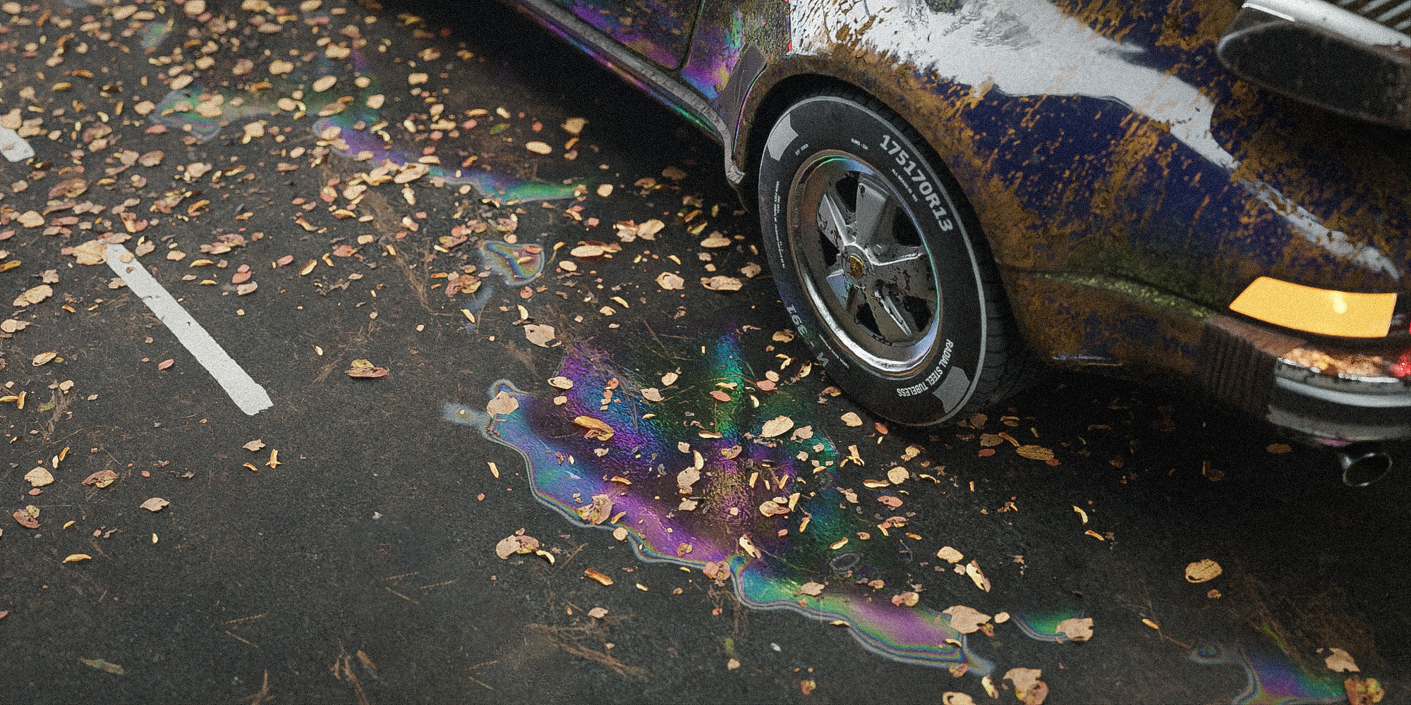 Iridescent oil stains on a road beside a weathered car.
